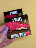 Sticker with a cartoon alligator and text 'I WILL KISS YOU' held by a hand on a yellow background