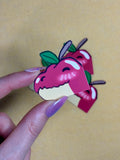 Hand holding a sticker in the shape of an alligator head with the colors of an apple above a yellow background.