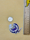 A sticker of the elden beast from elden ring in a chibi style next to a coin and ruler on a beige background