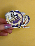 Hand holding a sticker of the elden beast from elden ring in a chibi style on a yellow background