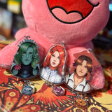 Characters from the Just Roll With It Podcast Riptide campaign as keychains. Gillian Tidestrider, Jay Ferin and Chip keychains are in front of the pink Pretzel plushie. 