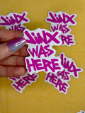 A hand holding a sticker that says JINX WAS HERE in pink text above a yellow background.
