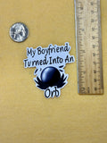 A sticker that reads 'my boyfriend turned into an orb' next to a ruler and a quarter on a yellow background